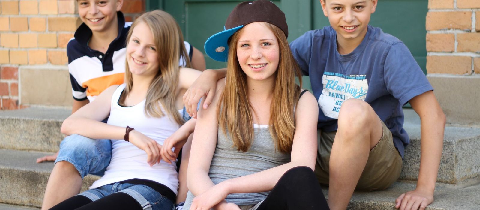 Vier Jugendliche (zwei Jungen, zwei Mädchen) sitzen auf einer Treppe vor einem Haus.