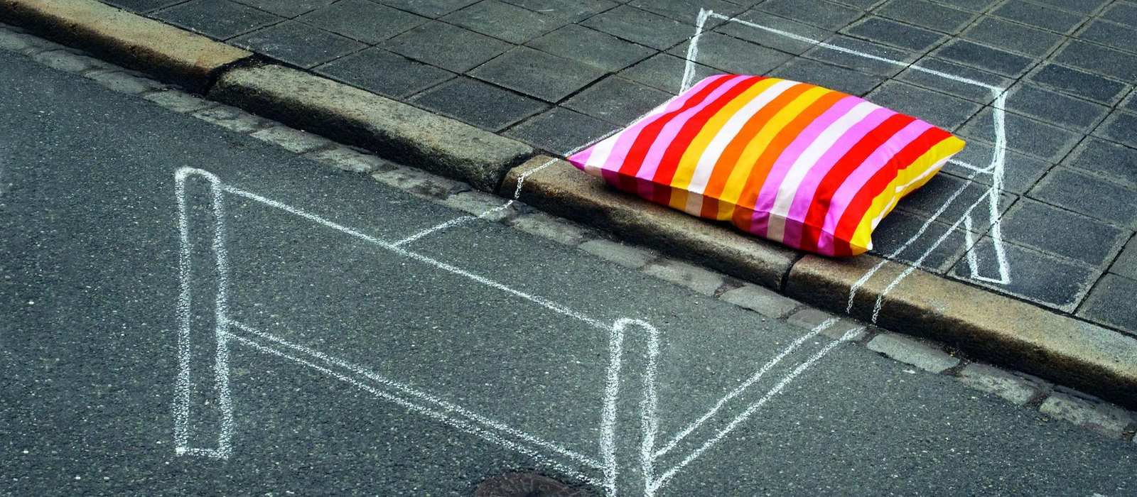 Auf eine Straße ist mit weißer Kreide ein Bett gezeichnet. Auf dem Kreidebett liegt ein bunt gestreiftes Kopfkissen.