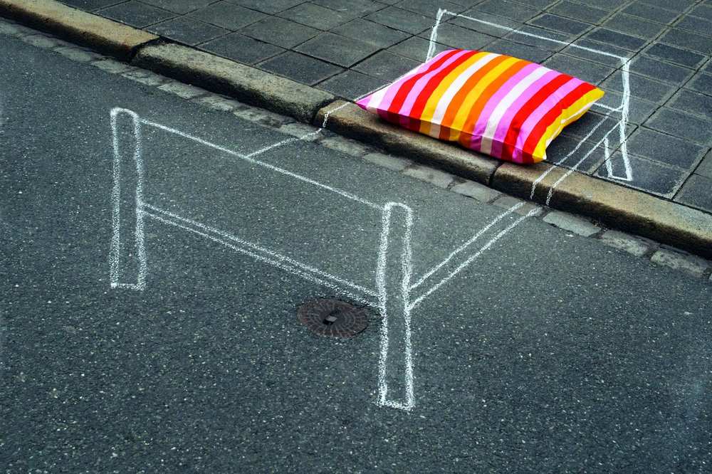 Auf eine Straße ist mit weißer Kreide ein Bett gezeichnet. Auf dem Kreidebett liegt ein bunt gestreiftes Kopfkissen.