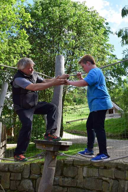 Ein junge im blauen T-Shirt balanciert über ein Seil. Ein Mann streckt ihm hilfreich die Hände entgegen.