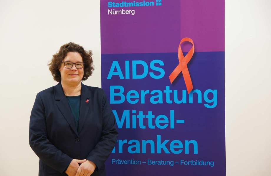 Eine Frau im dunkelblauen Sakko steht neben einem Roll-Up der AIDS-Beratung. Darauf prangt eine große rote AIDS-Schleife.