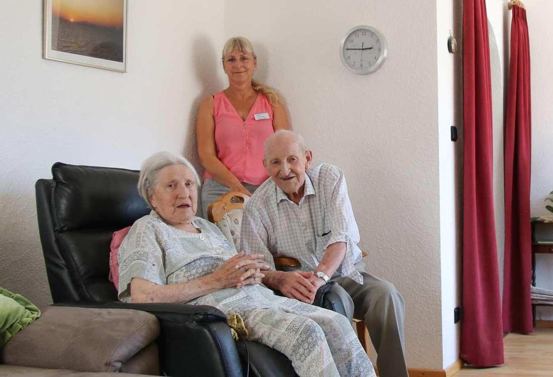 Ein älteres Ehepaar sitzt in ihrem Wohnzimmer. Hinter ihnen steht eine Pflegekraft.