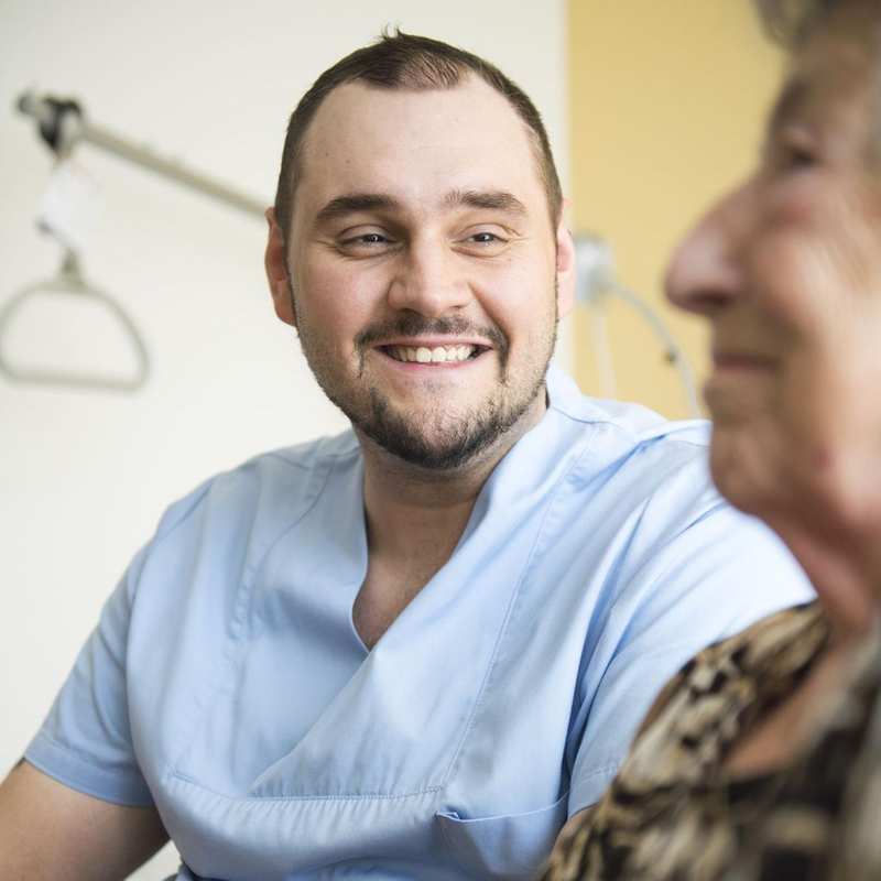 Ein Pfleger sitzt neben einer älteren Dame und lächelt.