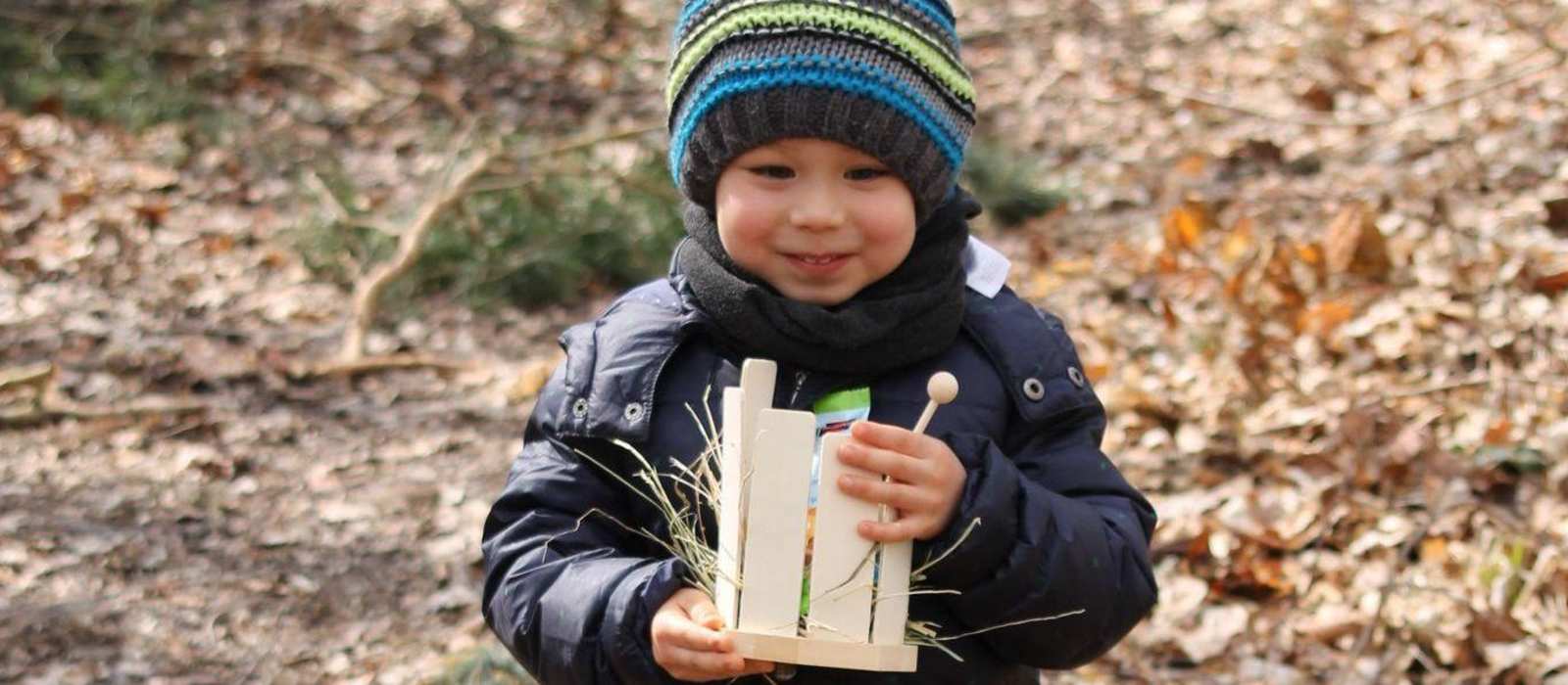 Ein warm gekleideter Junge trägt stolz sein Osternest durch den Garten mit Laub am Boden.