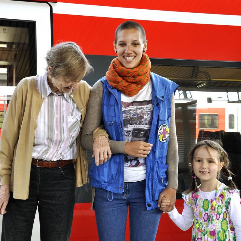 Eine alte Dame, eine junge Mitarbeiterin der Bahnhofsmission und ein kleines Mädchen stehen vor einem roten Zug. Die Dame henkelt sich bei der Helferin ein. Die Helferin hält das Mädchen an der Hand. 