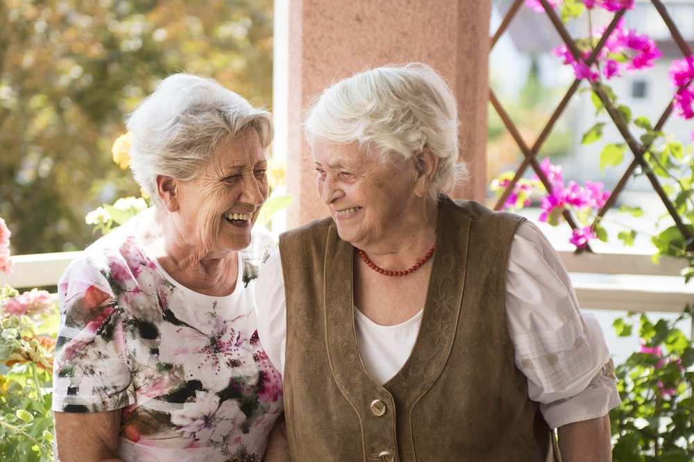 Zwei Seniorinnen stehen auf einem begrünten Balkon und lachen miteinander.