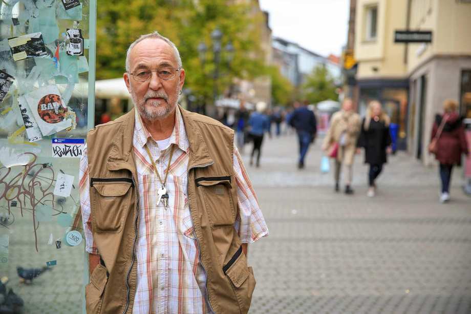 Ein älterer Mann mit weißem Haar, Brille und Dreitagebart in der Fußgängerzone. Er trägt ein kariertes Hemd und eine braune Weste.