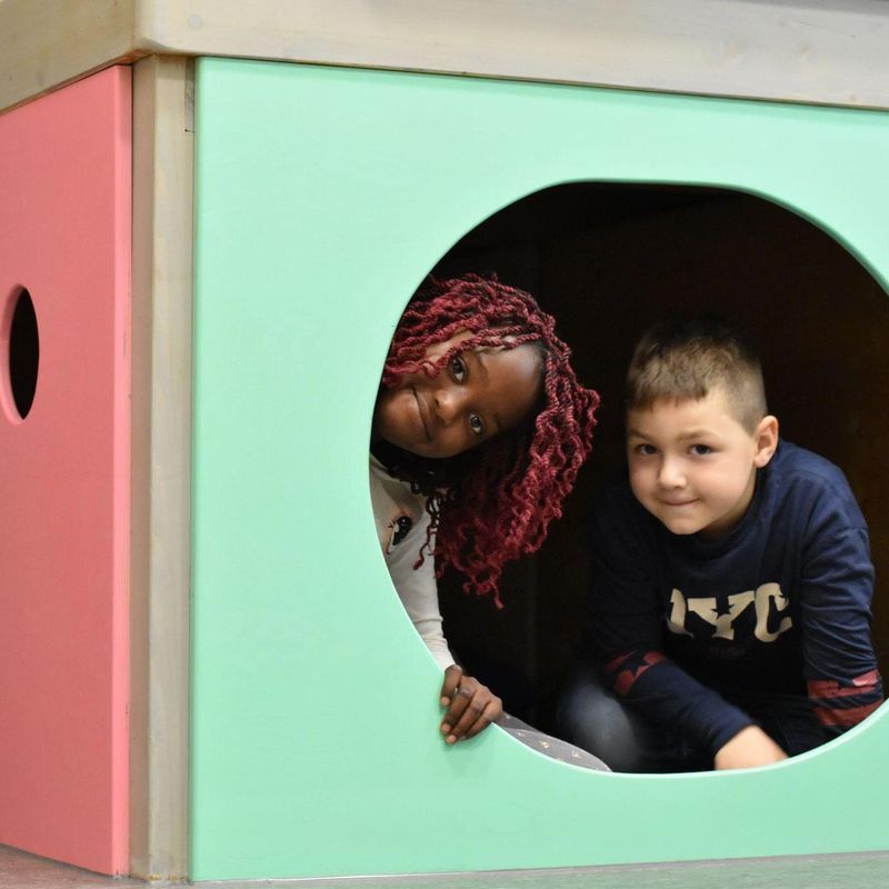 Zwei Kinder verstecken sich in einer Art Indoor-Höhle und schauen aus dem runden Loch.