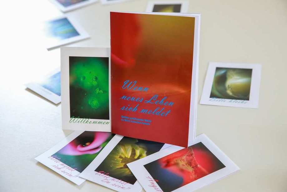 Auf einem Tisch liegen Karten mit bunten Fotografien. Dazwischen ein rotes Heft mit der Aufschrift "Wenn neues Leben sich meldet"