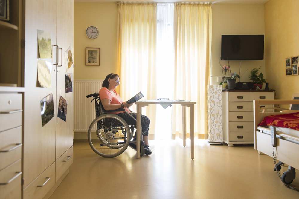 Eine Frau sitzt in ihrem Rollstuhl in ihrem Zimmer und liest Zeitung.