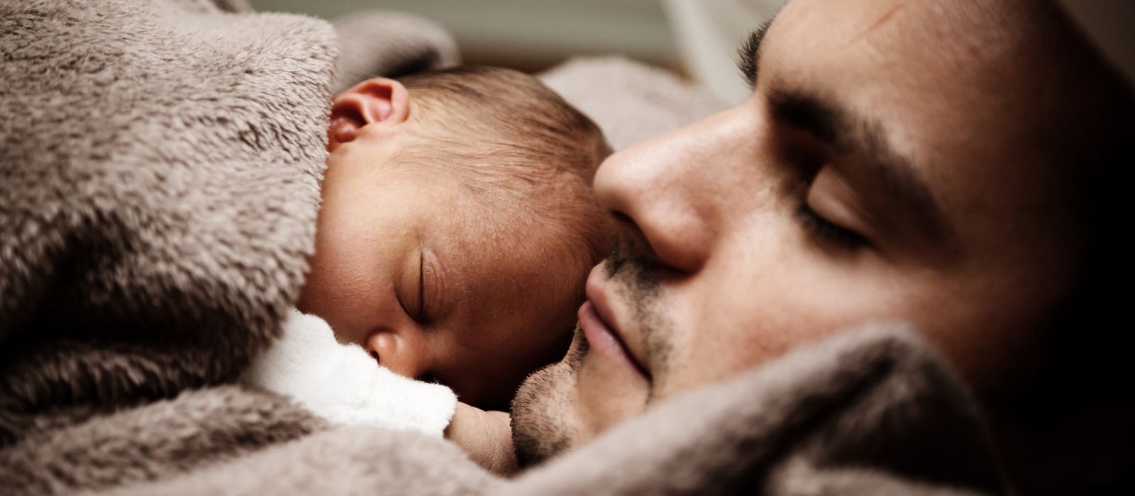 Ein Mann liegt mit einem Baby auf der Brust und mit einer Decke umkuschelt da. Beide haben die Augen geschlossen.
