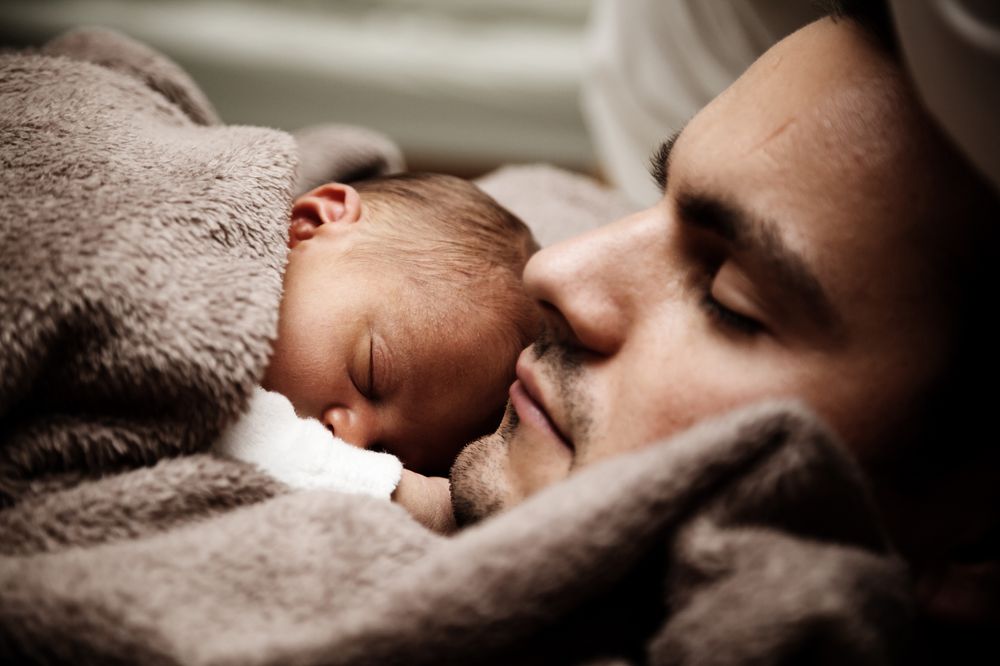 Ein Mann liegt mit einem Baby auf der Brust und mit einer Decke umkuschelt da. Beide haben die Augen geschlossen.