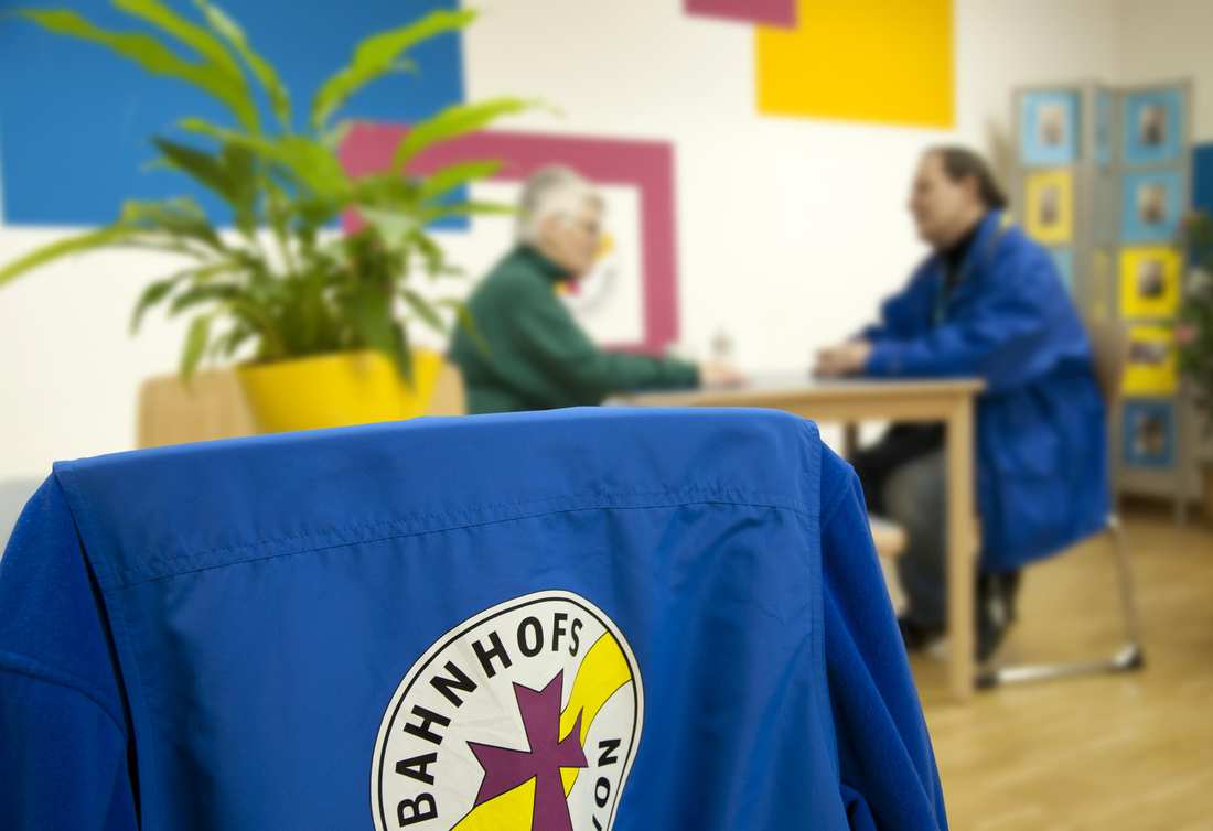 Im Vordergrund hängt eine blaue Bahnhofsmission-Jacke über einem Stuhl. Im Hintergrund ist ein Beratungsgespräch zu sehen.