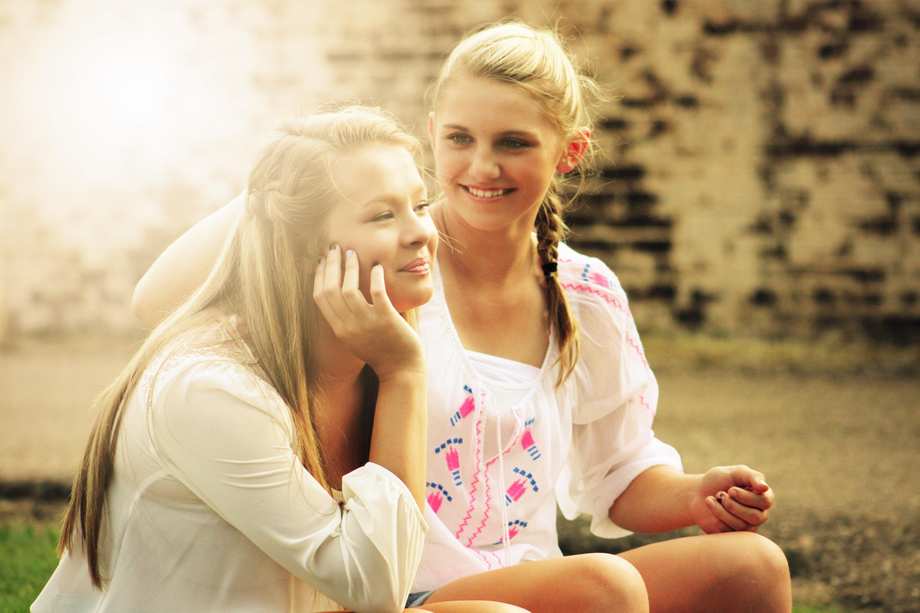 Zwei blonde Teenager-Mädchen sitzen an einer Mauer. Sie lächeln.