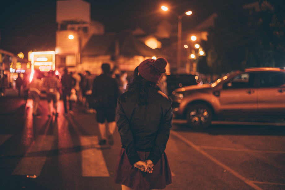Blick auf eine nächtliche Straße: Eine Frau hält die Hände hinter ihrem Rücken zusammen und läuft über eine Straße. Im Hintergrund sind andere Menschen unscharf zu erkennen.