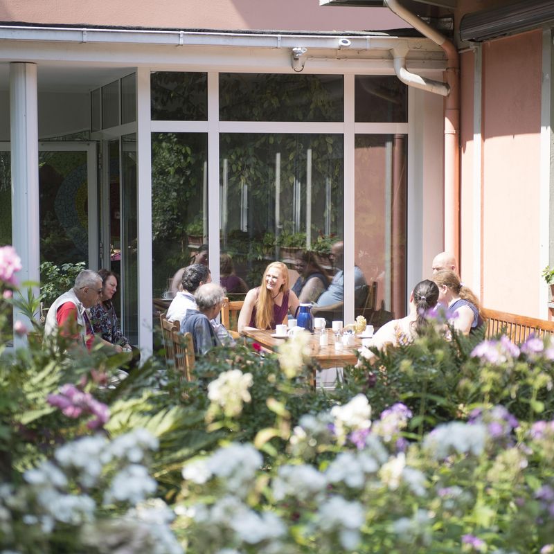 Eine Gruppe sitzt im sonnigen Garten vor einem Haus und unterhält sich.