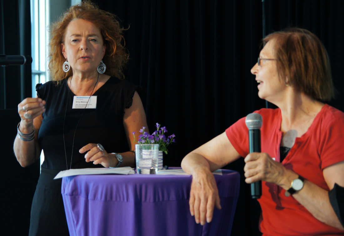 Zwei Frauen stehen mit Mikrofon an einem Stehtisch. Sie diskutieren.