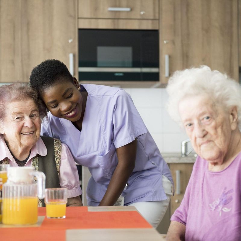 Eine Pflegerin steht bei zwei älteren Damen, die an einem Tisch sitzen.