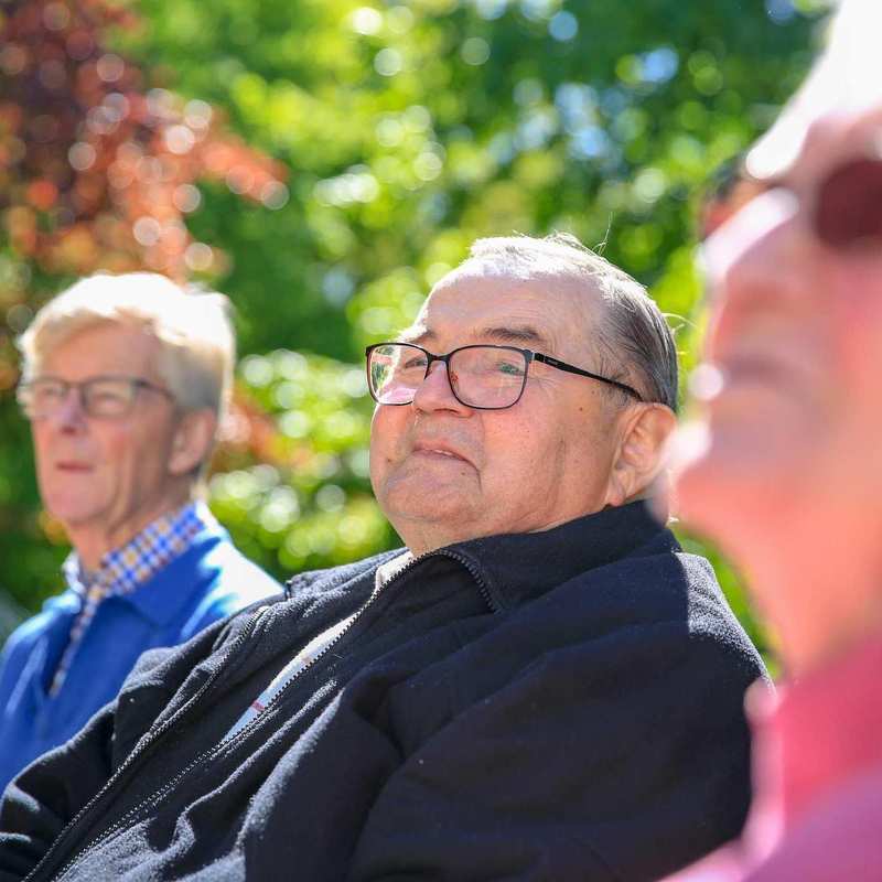 Drei ältere Personen mit Brillen sitzen zusammen auf einer Bank im Garten.