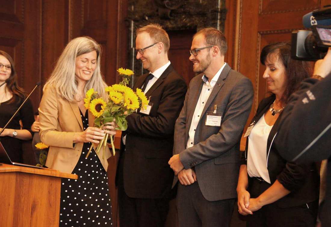 Eine Frau hält einen Strauß Sonnenblumen in der Hand. Sie verteilt diese an vier weitere Personen, die lächelnd vor ihr stehen.