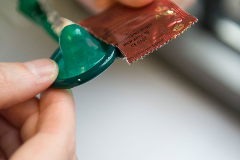 Zwei Hände ziehen ein grünes Kondom aus dessen roten Verpackung.