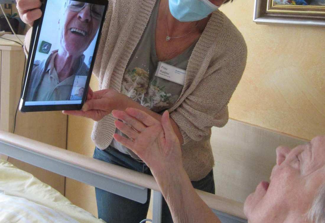 Eine Frau mit Mundschutz hält ein Tablet vor eine alte Dame, die in einem Pflegebett liegt. Auf dem Tablet-Bildschirm lacht ein älterer Herr.