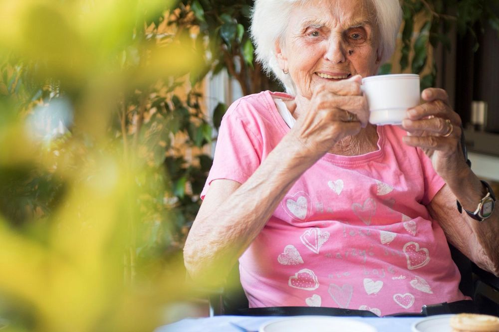 Eine ältere Dame sitzt im Grünen an einem gedeckten Tisch und hält eine Tasse in der Hand.