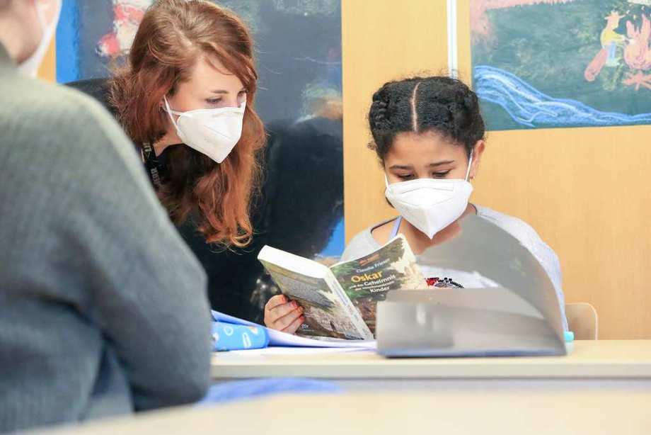An einem Tisch sitzt ein Mädchen mit einem Buch, daneben eine Erzieherin. Beide tragen Masken.