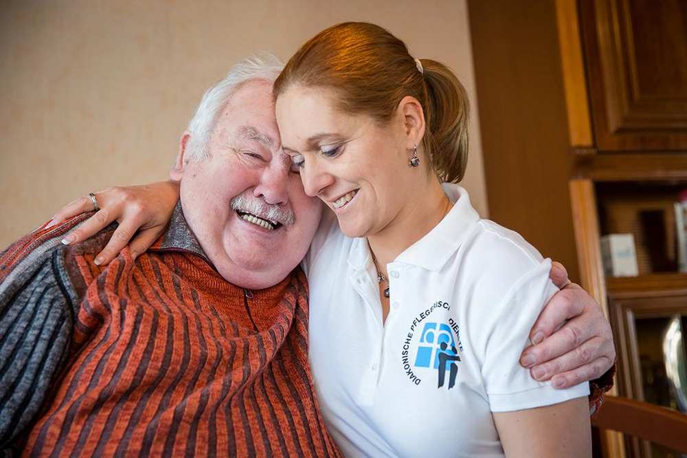Eine Pflegekraft und ein Senior liegen sich im Arm und lächeln.