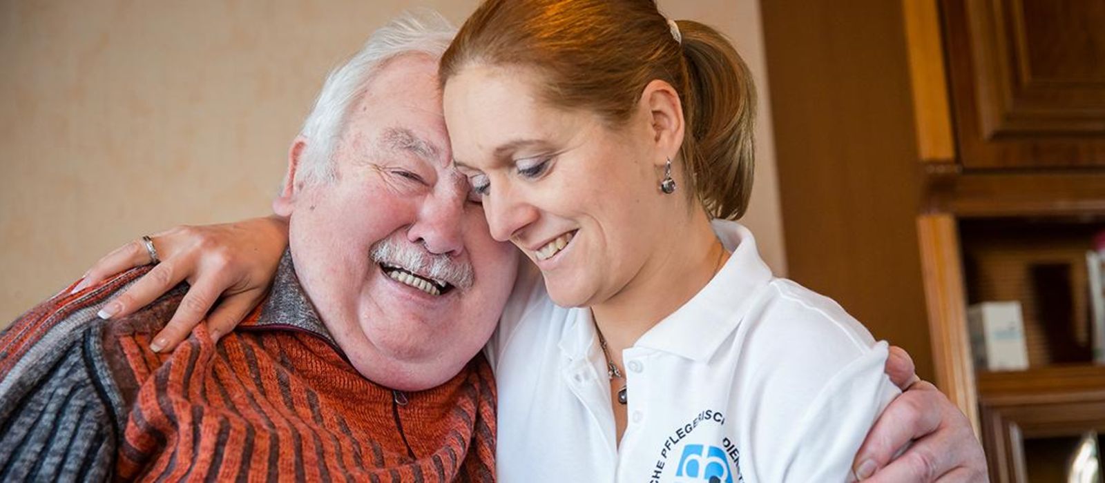 Eine Pflegekraft und ein Senior liegen sich im Arm und lächeln.