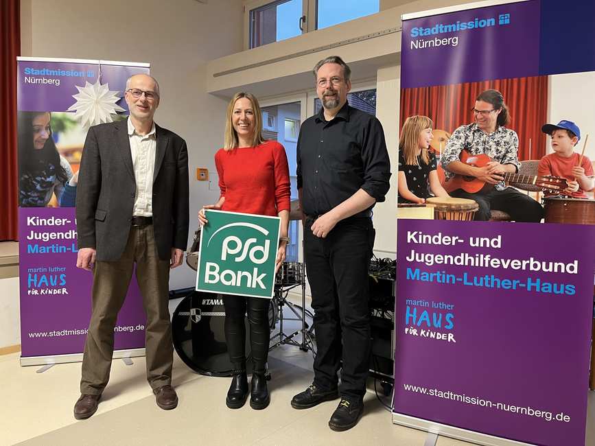 Zwei Männer und eine Frau stehen zwischen Plakaten der Stadtmission Nürnberg im Rahmen einer Spendenübergabe.