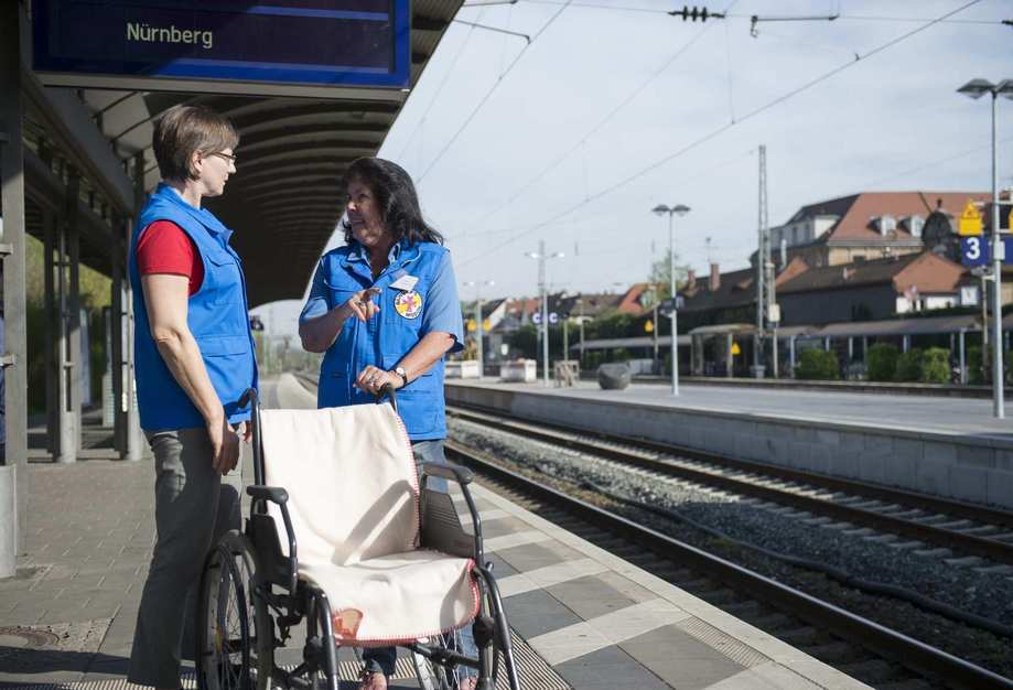Zwei Damen schieben einen Rollstuhl auf einem Bahnsteig.