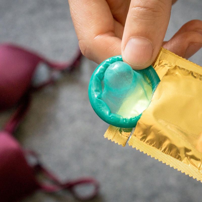 Ein Kondom wird aus der Verpackung gezogen, im Hintergrund liegt ein BH.