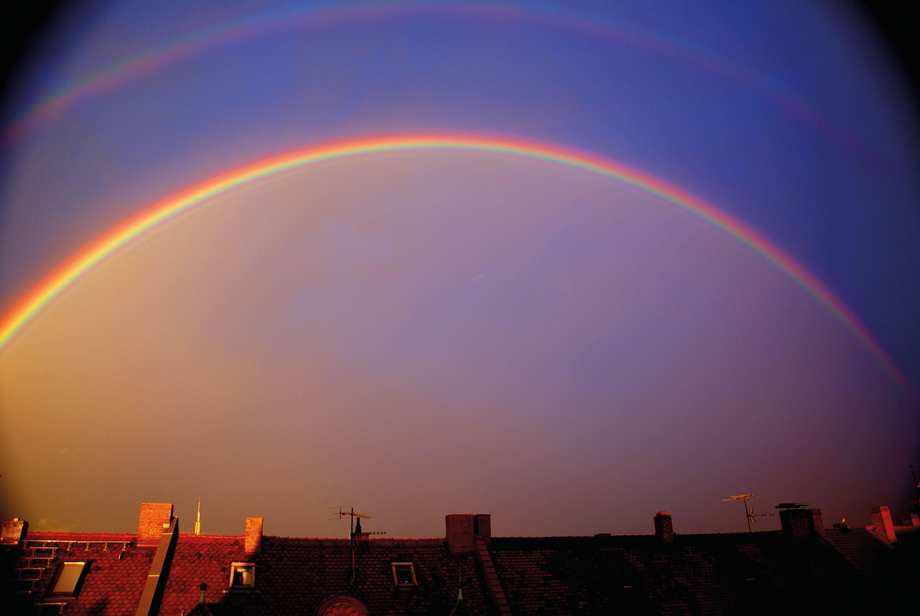 Ein Regenbogen steht über einer Dachsilhouette am dunkelblauen Himmel.